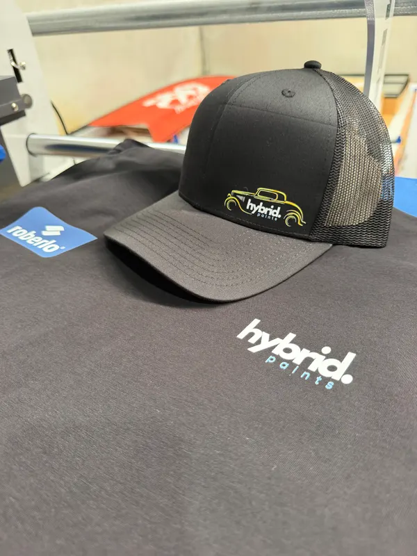 Black trucker cap with a stitched Hybrid Paints logo resting on a black tee printed with the Hybrid Paints logo